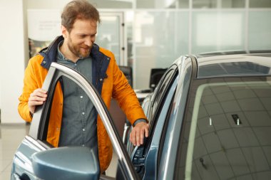 Güzel adam gülümsüyor ve araba merkezinde koyu mavi bir otomobilin yanında duruyor ve otomobili seçiyor. Modern araba galerisinde arabalara bakan yakışıklı bir erkek müşteri..