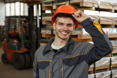 Portakal rengi kask takan ve metal kaskına dokunan mutlu erkek işçinin portresi. Gülümseyen adam gülüyor, poz veriyor ve kameraya bakıyor. Arka planda paletler ve forklift.