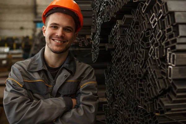 stock image Portrait of handsome smiling worker in orange hardhat and protective suit with folded arms standing near metal rods on metal base. Happy man looking at camera, posing and enjoying working process.