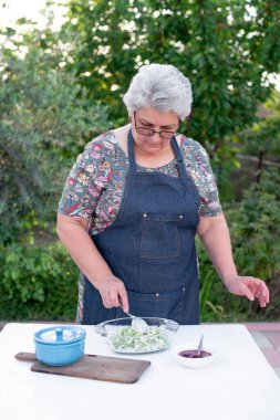 Önünde gri saçlı, önlük giyen, dışarıda yemek yapan, kaşığı tutan, salatayı karıştıran yaşlı bir kadın var. Yemek pişirme kavramı.