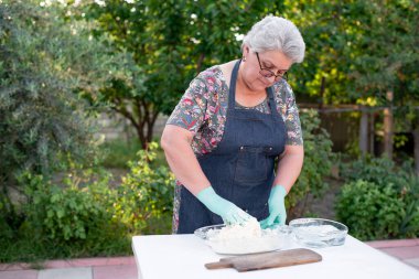 Gri saçlı, önlük takan, lateks eldivenli, aşağıya bakan, hamur yoğuran bir kadın. Yemek pişirme kavramı.