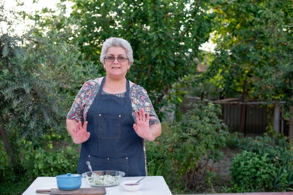 Gri saçlı, gözlüklü, dışarıda yemek pişiren, kameraya bakan, gülümseyen yaşlı bir kadın. Yemek pişirme kavramı.