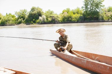 Nazik balıkçı, teknede oturup balık tutarken itaatkar bir köpeği evcilleştiriyor. Arka planda aşırı büyümüş bir bankayla elinde olta tutarken tüylü evcil hayvana sarılan nazik adamın ön görüntüsü. Arkadaşlık kavramı.
