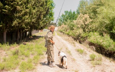 Kamuflaj kıyafetli dalgın bir balıkçı, güneşli bir günde sadık bir köpekle toprak yolda yürüyor. Çalı yolu boyunca balık tutmaya giderken sıska bir adamın elinde olta taşıyan omzundan bak. Yaşam tarzı kavramı.