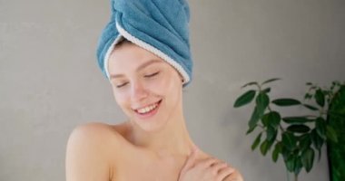 Duştan sonra genç ve hayalperest bir kadın, berrak cildine dokunuyor ve hareketli bir şekilde gülümsüyor. Banyoda gri arka planda mavi havluyla poz veren güzel bir kadın portresi. Güzellik bakımı kavramı.