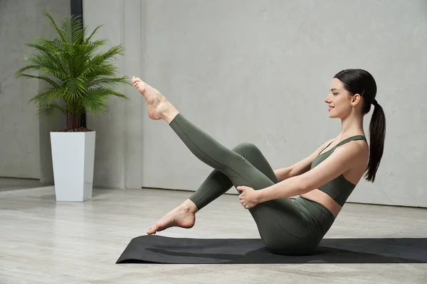 Stock image Side view of brunette girl wearing sportswear, training, exercising, working out. Attractive, young lady sitting barefoot on yoga mat, raising legs. Concept of working out and sport.