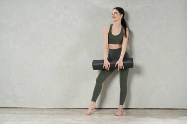 Çıplak ayaklı kadın spor giyer, spor yapar, spor yapar, ara verir. Genç, sıska bir kadın, yana bakıyor, gülümsüyor, yoga minderini tutuyor. Egzersiz ve eğitim kavramı.