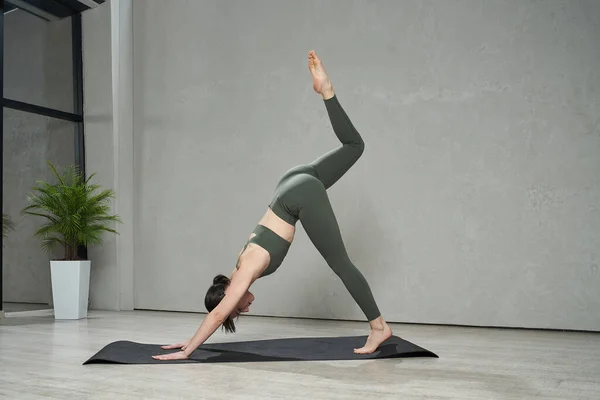 stock image Side view of flexible, active lady exercising, working out, practising in studio. Beautiful, slim girl standing barefoot on yoga mat, strtching. Concept of sport and wellbeing.