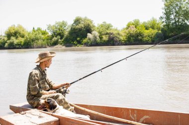 Genç, odaklanmış, kamuflajlı bir adam balıkçı teknesinde kıç tarafında, iskeleye demirlemiş olarak otururken. Balıkçı erkeğin olta tutarken yandan görünüşü, yorgun köpeğin dinlenmesi, derin nefes alması. Hobi kavramı.