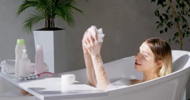 Hafta sonları küvette dinlenirken kollarına köpük saçan güzel bir kadın. Modern banyoda banyo yaparken süngerden köpük sıkan mutlu kadın modelin yan görüntüsü. Vücut bakımı kavramı.