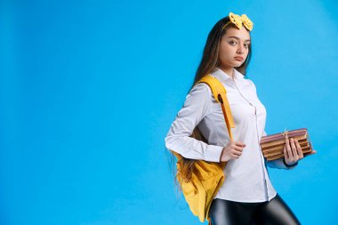 Sırt çantalı düşünceli, ciddi bir kız elinde bir sürü kitapla, hüzünle kameraya bakıyor. Puantiyeli saç bandı takan kendine güvenen kız öğrencinin portresi, mavi üzerine izole edilmiş. El kol hareketi kavramı.
