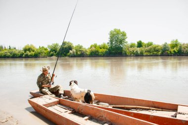 Odaklanmış erkek balıkçı, kıyıda eski püskü bir teknede otururken çelme takıyor. Balıkçının sevimli köpeğiyle panoramik manzarası, balıkçılık, arka planda sakin bir nehir yayılması. Hobi kavramı.