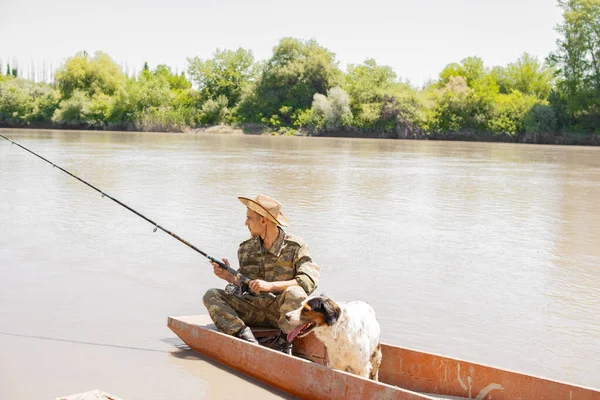 Relaxed angler in camouflage holding fishing pole, resting, while floating on lake with dog on board. Side view of pensive male fishing from boat, with landscape on background. Concept of fishery.