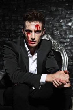 Kanepede oturan yakışıklı bir gencin portresi. Takım elbise giymiş, yüzünde kanlı bir makyajla elleri çaprazlanmış, ciddi görünüyor.