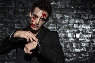 Ayakta duran ve saatlerine bakan siyah takım elbiseli, yüzünde kan olan bir zombi işadamının portresi.