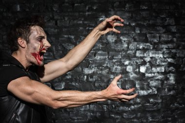 Kanlı makyajlı genç bir adamın belden yukarı fotoğrafı, bir profilin içinde durmuş bağırıyor ve siyah tuğla duvardaki yeri kopyalamak için ellerini uzatıyor.