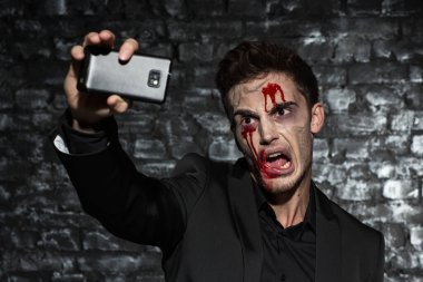Genç bir adamın portresi, Cadılar Bayramı için kanlı makyajlı siyah bir takım elbise giyiyor, akıllı telefonunda korkunç yüzüyle selfie çekiyor.
