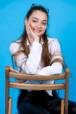 Gülümseyen, dalgalı sarı saçlı ve şehvetli bakışlı genç bir kadının portresi. Sandalyeye oturup mavi stüdyo arka planında izole edilmiş kameraya bakarken. Poz verme kavramı.