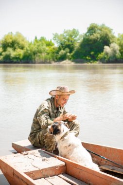 Odaklanmış gülümseyen erkek balıkçı, gölde eski püskü bir teknede dinlenirken oltaya yem takıyor. Yaz tatili boyunca sevimli bir köpekle balığa çıkan neşeli balıkçıların ön görüntüsü. Hobi anlayışı, hobi anlayışı.