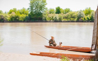 Balıkçılık sırasında, iskelede köpekli bulutlu suda yüzen şapkalı bir balıkçı. Arka planda çalılık kıyıları ile, dinlenirken, sakin erkek bakış açısı. Hobi kavramı.