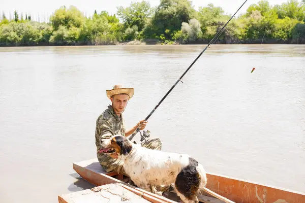 Young fisherman in hat holding fishing pole, while sitting in rowing old boat at pier in morning. Side view of caring man in camo looking at thirsty dog, while angling in calm river. Concept of rest.