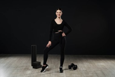 Stüdyoda spor malzemeleriyle poz veren aktif giyimli güzel bir kadın. Kameraya gülümseyen ince yoga eğitmeninin düşük açılı görüntüsü, siyah duvarın karşısında dikilirken, fotokopici. Spor anlayışı.