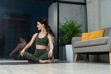 Çekici beyaz kadın modern dairede Sukhasana pozisyonunda yoga minderinde oturuyor. Düşük açılı mutlu, sakin esmer akşam mesaisinden sonra rahat bir poz veriyor. Yoga, meditasyon konsepti..