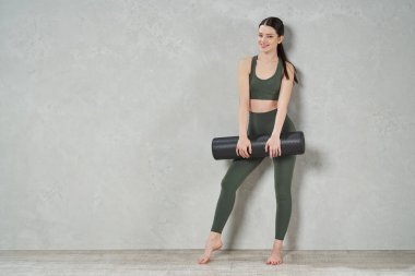 Stüdyoda yoga minderiyle poz veren genç ve güzel bir kadın. Aktif giysi giyen, kameraya gülümseyen, gri duvarlı, fotokopici kadının düşük açılı görüntüsü. Spor konsepti.