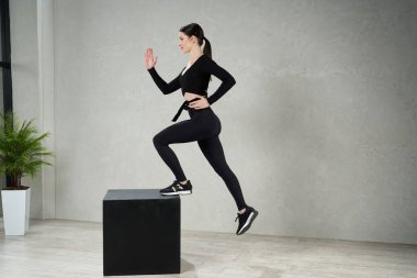 Siyah küpün üzerine atlayan ve gri arka planda egzersiz yapan genç bayan sporcu. Spor ayakkabılı güçlü bir kadının modern spor salonunda çalışırken tahta küpün üzerine basması. Eylem kavramı. 