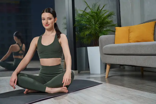 Peaceful beautiful female sitting in Sukhasana position on yoga mat near sofa at home. Front view of calm brunette woman in sports bra relaxing in easy yoga pose in modern bedroom. Yoga concept.