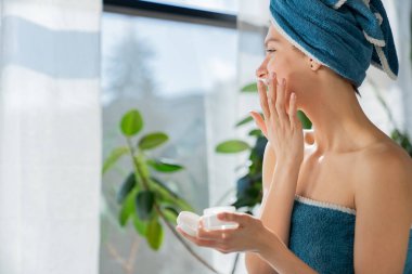 Kafasında havlu olan beyaz bir kadın duştan sonra yüz kremi sürüyor, pencereden bakıyor. Hafif banyoda yüz kremi tutan mutlu bir kızın yan görüntüsü. Cilt rutini kavramı.