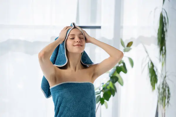 stock image Beautiful young female wrapping hair in turquoise towel after showering in light room. Portrait of happy, relaxed woman putting towel on wet head in bathroom. Concept of hair care routine, beauty.