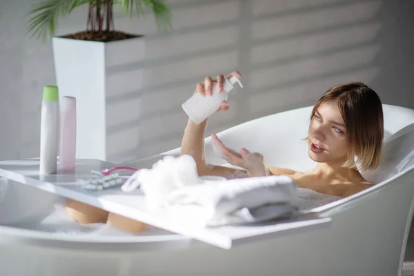 Beautiful woman pouring body wash on hand, while resting in bathtub. Side view of sexy female taking bath, with set of bath accessories on bath table, in sunlit bathroom. Hygiene, cosmetics concept.