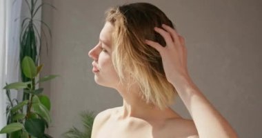 Göz kamaştırıcı genç bir kadın saçlarını kurutuyor ve pencereden banyoya bakıyor. Sarışın, çıplak omuzlu, kısa, havadar saçlara dokunan, güneşli bir sabahta kuruyan kadının yan görüntüsü. Yaşam tarzı kavramı.