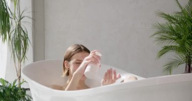 Genç sarışın kadın küvette otururken sıvı sabun sıkıştırıyor. Baştan çıkarıcı bir kadının, otelde banyo yaparken palmiyeye dökülen vücudunun yan görüntüsü. Kişisel bakım, kaplıca, hijyen konsepti.