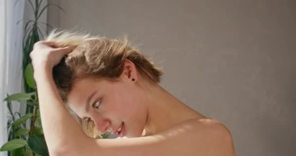魅力的な若い女性は 浴室の窓を見ながら 乾いた髪を吹き飛ばします 裸の肩を乾燥させ 朝に短い染められた髪に触れる白人女性の側面の景色 日々のルーチンコンセプト — ストック動画