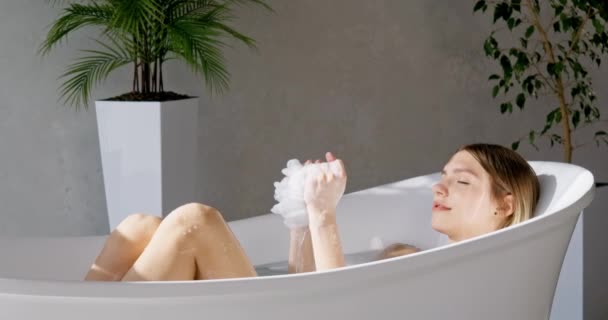 スタイリッシュなバスタブで浸っている間 スポンジで洗う穏やかな美しい女性 リラックスしたブロンドの女性の絞りスポンジのサイドビューは 豪華なホテルで風呂に入りながら ゆっくりと動きます リラックス スパコンセプト — ストック動画