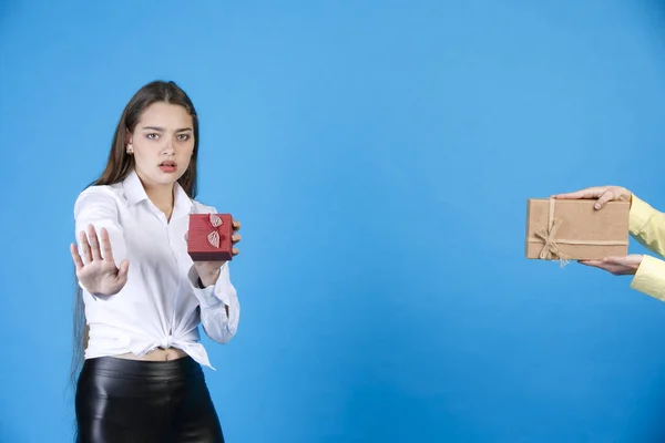 Discontented woman with small box holding arm out by refusing gift. Front view of disgruntled lady making stop gesture against camera, with crop hand with gift, isolated on blue. Concept of gesturing.