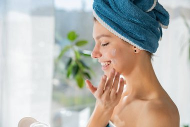 Çıplak omuzlu mutlu bir kadın, banyoda duştan sonra yüz kremi kullanıyor. Kapalı gözlü neşeli bir kadın yüzü nemlendirici, güneşli pencereye karşı. Cilt bakımı kavramı.