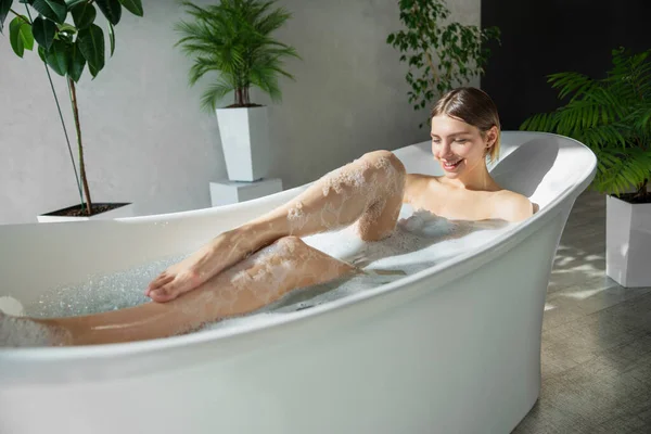 Happy female with haircut, washing body in bubble bathtub in sunny morning. Side view of carefree, relaxed girl with bare shoulders soaking in bath, surrounded with plants. Bathing, rest concept.