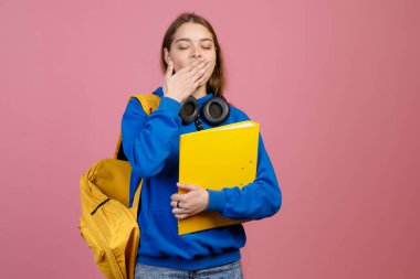 Güzel liseli kızın gözleri kapalı, esniyor. Mavi khudi ve kot pantolon giyen, klasör ve sırt çantası tutan zayıf bir kadın. Üniversite ve eğitim kavramı.