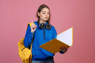 Mavi khudi ve kot pantolon giyen, kitap okuyan güzel okul kızlarının ön görüntüsü. Genç kız öğrenci dosyasını açık tutuyor, parmağını kaldırıyor, dikkat çekiyor. Öğrenci hayatı kavramı.