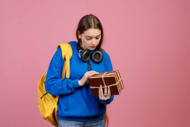 Elinde bir sürü eski kitapla ayakta duran esmer güzel bir kız. Güzel liseli kız ders çalışıyor, aşağı bakıyor, sarı sırt çantasıyla. Pembe stüdyo arka planında izole edilmiş..