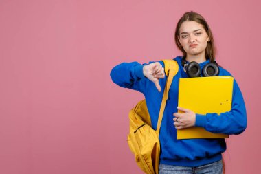 Genç bir kız öğrencinin ayakta durması hoşnutsuzluk göstergesidir. Güzel kız kameraya bakıyor, kasvetli, sarı sırt çantası ve dosya tutuyor, okuldan nefret ediyor. Gençlik kavramı.