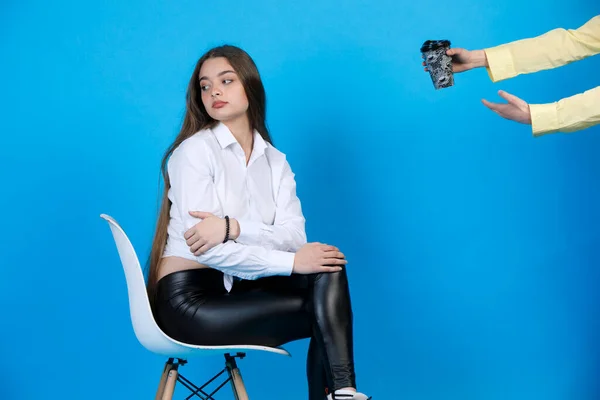 Upset, offended brunette lady in leather leggings turning away, while unrecognisable hands in yellow shirt offering paper cup of coffee, isolated on blue studio background. Concept of resentment.