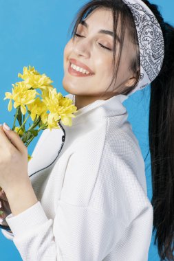 Sarı kasımpatı ile poz veren genç, esmer bir kadın, kokuyor. Mavi stüdyo arka planında izole edilmiş..