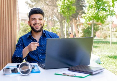 Masada oturan, çalışan, dizüstü bilgisayar kullanan yakışıklı genç girişimcinin ön görüntüsü. Sakallı esmer çocuk kameraya bakıyor, gülüyor, mavi gömlek giyiyor. Gençlik kavramı.