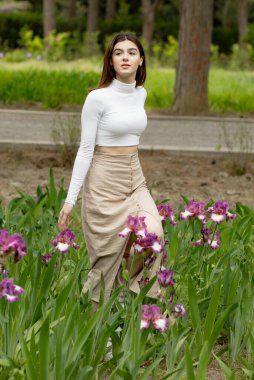 Çekici, zayıf, esmer bir kadın bahçede çiçeklerle yürüyor, yan bakıyor, eğleniyor. İlkbahar kavramı.