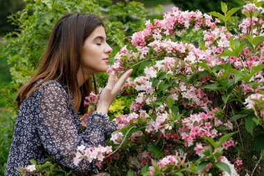 Güzel, esmer bir kızın çiçek açan ağacın yanında dikilip eğilmesinin, pembe çiçeklerin kokusunu almasının yan görüntüsü. İlkbahar kavramı.