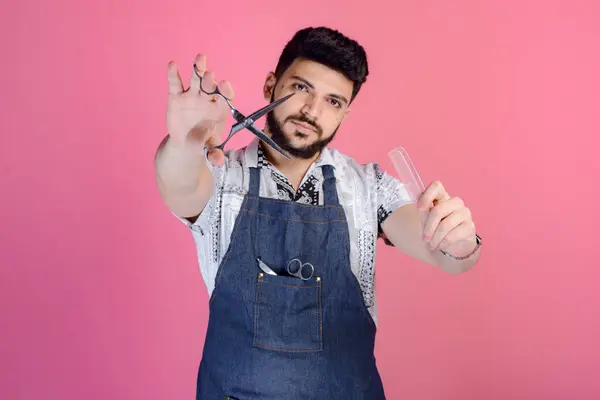Front view of male hairdresser, barber standing, looking at camera, showing scissors and comb. Isolated on pink studio background.